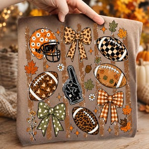 May include: A brown t-shirt featuring a graphic print of football-themed elements including an orange daisy-patterned helmet, a black foam finger, four decorative bows, and four footballs with various patterns like leopard print, hearts, and checkers.