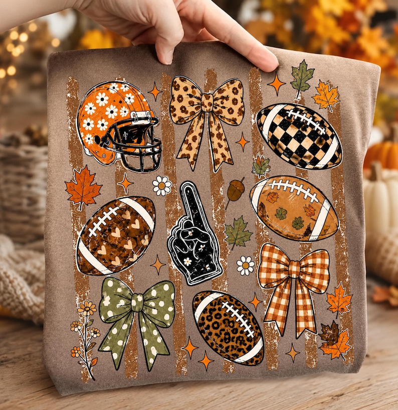 May include: A brown t-shirt featuring a graphic print of football-themed elements including an orange daisy-patterned helmet, a black foam finger, four decorative bows, and four footballs with various patterns like leopard print, hearts, and checkers.