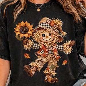May include: A charcoal black t-shirt featuring a graphic of a scarecrow holding a large yellow sunflower. The scarecrow wears a hat with a black and white checkered pattern, a vest, and boots with leopard print and plaid accents.