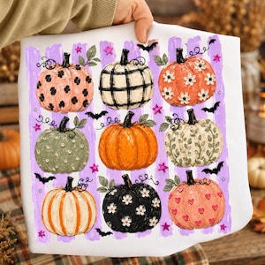 May include: A white t-shirt featuring a graphic print of nine pumpkins arranged in a grid. The pumpkins display various patterns including polka dots, plaid, floral motifs, and hearts, set against a background of vertical purple brush strokes.