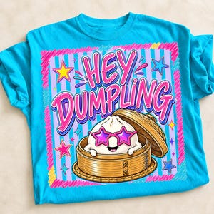 Może przedstawiać: Turkusowa koszulka z grafiką pierożka w okularach przeciwsłonecznych w kształcie gwiazd w bambusowym parowarze. Tekst "HEY DUMPLING" jest w kolorze różowym i białym, obrysowany na czarno. Projekt jest obramowany różowymi i fioletowymi paskami i gwiazdami.
