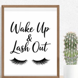May include: A framed print with the words "Wake Up & Lash Out" in black script, along with a pair of stylized eyelashes. The frame is a light brown color. A cactus in a white pot is to the right of the frame.