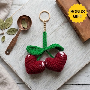 May include: A crochet keychain featuring two dark red heart-shaped cherries with white highlights, connected by green crocheted stems and leaves to a gold-toned metal split ring. The cherries are textured with yarn stitches and have a soft appearance.
