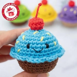 May include: A handmade, blue crochet cupcake with a red cherry on top. The cupcake has a brown base, yellow sprinkles, and a smiling face. Other colorful cupcakes are in the background. The image includes a "BONUS GIFT INCLUDED" logo.