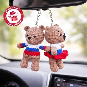 May include: Two crocheted teddy bear car charms. The bears are brown with white, red, and blue accents. One bear has a skirt, and the other has a shirt with matching colors. They hang from a silver chain.