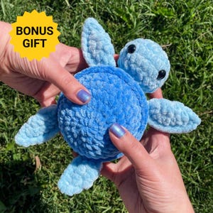 Peut inclure: Une peluche tortue en crochet bleu clair avec une finition en fil doux et texturé, comprenant une carapace ronde, quatre nageoires et une tête avec deux yeux en plastique noir. Un graphique en étoile jaune affiche le texte BONUS GIFT.