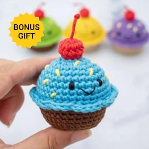 May include: A small blue crocheted cupcake toy with a brown base, yellow sprinkles, and a red cherry on top. The cupcake features a stitched smiling face with black eyes and a curved mouth. Additional crocheted cupcakes in green, yellow, and purple are visible in the background with a yellow starburst graphic labeled BONUS GIFT.