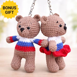 May include: Two brown crocheted bear keychains with metal chains. The left bear wears a white, blue, and red striped sweater. The right bear wears a white and blue top with a red ruffled skirt. A yellow star-shaped badge says BONUS GIFT.