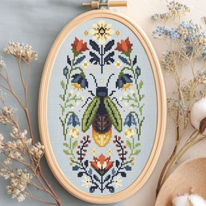 May include: An oval wooden embroidery hoop containing a cross-stitch pattern of a central firefly with green wings and a yellow abdomen, surrounded by symmetrical floral and botanical motifs in shades of red, blue, green, and white.