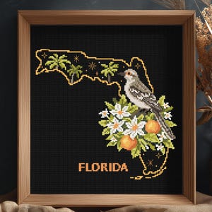 May include: A framed cross-stitch embroidery featuring the outline of Florida in yellow on a black background. The design includes palm trees, a mockingbird, orange blossoms, and oranges, with the word FLORIDA printed in yellow at the bottom.