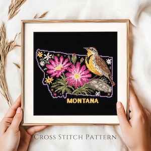 Puede incluir: Un patrón de punto de cruz enmarcado que representa el estado de Montana en negro, con flores rosas, un pájaro y la palabra "MONTANA" en oro. El marco es dorado y el fondo es de tela blanca.