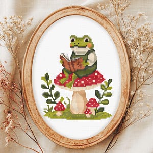 May include: Cross-stitch art of a green frog wearing a waistcoat and reading a book while sitting on a red mushroom with white spots. The artwork is framed in an oval wooden hoop, with green foliage and small mushrooms around the base.