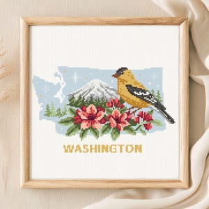 May include: A square cross-stitch embroidery in a light wood frame featuring the state of Washington. The design includes a yellow goldfinch, a snow-capped mountain, green pine trees, and pink rhododendron flowers, with the text WASHINGTON below.