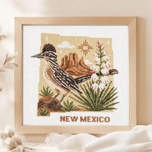 May include: A framed cross-stitch embroidery featuring a roadrunner, desert landscape, yucca plant, and the Zia sun symbol set against a tan map outline of New Mexico. The text NEW MEXICO is stitched in brown at the bottom of the white fabric.