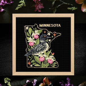 May include: A framed cross-stitch featuring the outline of Minnesota, with the state name in gold. The design includes a loon, pink flowers, and green foliage on a black background. The frame is light wood.