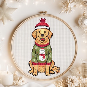 May include: A cross-stitch pattern in a wooden embroidery hoop featuring a golden retriever dog wearing a red knit hat and a green holiday sweater with a white snowman graphic. The dog has yellow fur, a black nose, and a happy expression.