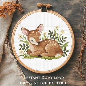 May include: A cross-stitch pattern of a sleeping fawn surrounded by flowers and greenery, set within a wooden embroidery hoop. The fawn is brown with white spots. The text "INSTANT DOWNLOAD CROSS STITCH PATTERN" is at the bottom.