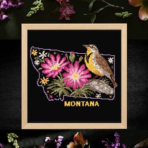Puede incluir: Un bordado a punto de cruz enmarcado que representa Montana, con flores rosas, un pájaro y la palabra "MONTANA" en dorado. El fondo es negro y el marco es de color marrón claro. Acentos florales rodean el marco.