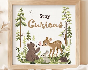 May include: A square cross-stitch pattern in a light wood frame featuring a forest scene with a brown bear, a fawn, a rabbit, and a hedgehog. The text "Stay Curious" is centered at the top in brown letters, surrounded by green pine trees.