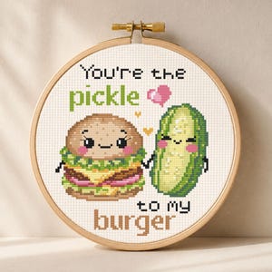 Puede incluir: Arte de aro de punto de cruz con una hamburguesa y un pepinillo sonrientes, con el texto "You're the pickle to my burger". El diseño incluye un corazón rosa y está en un aro de bordado de madera.