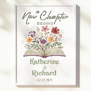 May include: A framed cross-stitch design featuring the words "New Chapter Begins" above a floral arrangement and an open book. Below, it reads "Katherine & Richard 12.27.2025". The design is in a white frame.