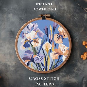 May include: A cross-stitch pattern of irises in a wooden embroidery hoop. The design features blue, purple, and peach-colored flowers against a light blue background. The text "INSTANT DOWNLOAD" and "CROSS STITCH PATTERN" are visible.