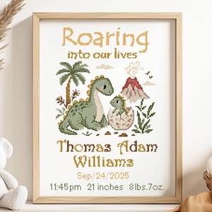 May include: A framed cross-stitch art piece with a dinosaur theme. The artwork features a green dinosaur, a baby dinosaur hatching from an egg, a palm tree, and a volcano. Text includes the phrase "Roaring into our lives" and the name "Thomas Adam Williams". The frame is 21 inches.