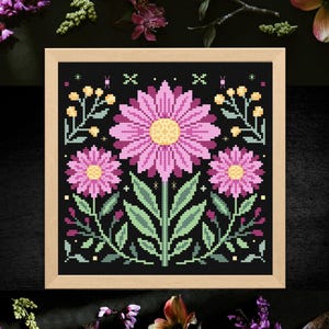 May include: A framed cross-stitch floral design featuring three pink flowers with yellow centers on a black background. The design includes green leaves and stems, with a light wood frame. Decorative floral elements surround the frame.