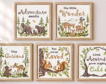 May include: Five square cross stitch patterns in light wood frames featuring woodland themes. Designs include bears, deer, squirrels, and trees with text: Adventure awaits, Our little Wonder, Stay Curious, You are so Loved, and Little Acorns grow mighty Oaks.
