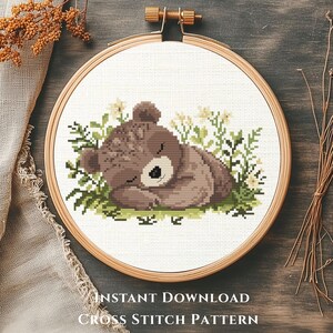 May include: A cross-stitch pattern of a sleeping brown bear surrounded by green foliage and small white flowers, displayed in a wooden embroidery hoop. The text "INSTANT DOWNLOAD CROSS STITCH PATTERN" is visible.