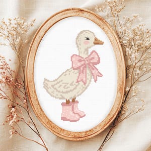 Può includere: Un disegno a punto croce di un'anatra con stivali rosa e un fiocco rosa. L'anatra è all'interno di una cornice ovale in legno, circondata da fiori secchi. L'anatra è color crema con un becco marrone.