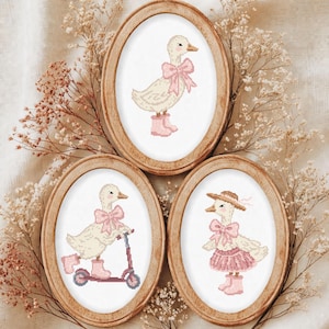 Könnte beinhalten: Drei ovale Holzrahmen mit Kreuzstich-Stickereien. Jedes Bild zeigt eine cremefarbene Ente mit rosa Akzenten. Eine Ente trägt eine Schleife und Stiefel, eine andere fährt einen rosa Roller und die dritte trägt einen Hut, eine Schleife und einen Rock.