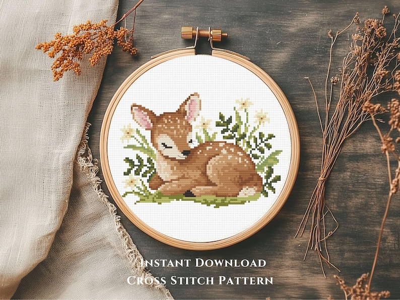 May include: A cross-stitch pattern of a sleeping fawn surrounded by flowers and greenery, set within a wooden embroidery hoop. The fawn is brown with white spots. The text "INSTANT DOWNLOAD CROSS STITCH PATTERN" is at the bottom.