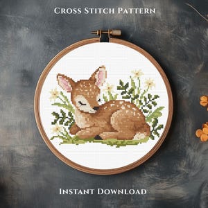 May include: A cross-stitch pattern featuring a fawn lying in grass and flowers, framed in a wooden embroidery hoop. The fawn is brown with white spots and pink ears. The text "CROSS STITCH PATTERN" and "INSTANT DOWNLOAD" are at the top and bottom.