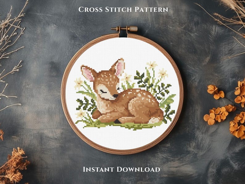 May include: A cross-stitch pattern featuring a fawn lying in grass and flowers, framed in a wooden embroidery hoop. The fawn is brown with white spots and pink ears. The text "CROSS STITCH PATTERN" and "INSTANT DOWNLOAD" are at the top and bottom.