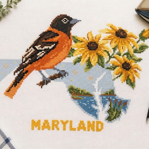 May include: A cross-stitch embroidery on white fabric featuring a Baltimore oriole bird, yellow sunflowers, and a light blue map outline of Maryland. The word MARYLAND is stitched in yellow letters below the map, accompanied by a small flag.