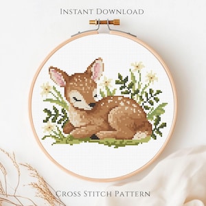 May include: Cross-stitch pattern of a fawn lying in grass and flowers, framed in a wooden hoop. The fawn is brown with white spots. The text "INSTANT DOWNLOAD" and "CROSS STITCH PATTERN" are at the top and bottom of the hoop.