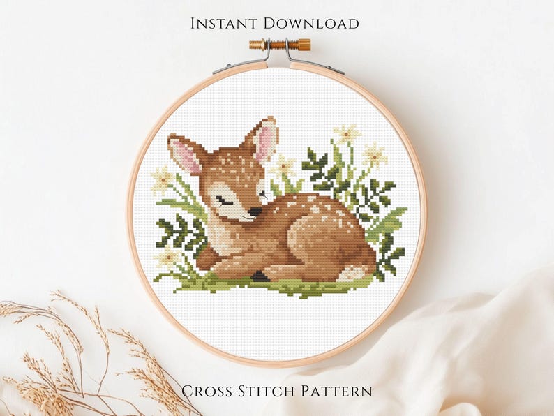 May include: Cross-stitch pattern of a fawn lying in grass and flowers, framed in a wooden hoop. The fawn is brown with white spots. The text "INSTANT DOWNLOAD" and "CROSS STITCH PATTERN" are at the top and bottom of the hoop.