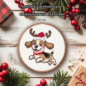 May include: A cross stitch pattern in a wooden embroidery hoop featuring a cartoon beagle dog wearing a red and white Santa hat with reindeer antlers. The text INSTANT DOWNLOAD CROSS STITCH PATTERN is printed above the hoop on a white surface.