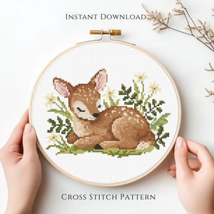May include: A cross stitch pattern of a sleeping fawn surrounded by flowers and greenery, set within a wooden embroidery hoop. The text "INSTANT DOWNLOAD" and "CROSS STITCH PATTERN" are visible.