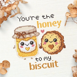 May include: Cross-stitch design featuring a jar of honey and a biscuit, each with smiling faces and hearts. The text reads "You're the honey to my biscuit." The design is in shades of yellow, brown, and white, with decorative dried flowers in the corners.