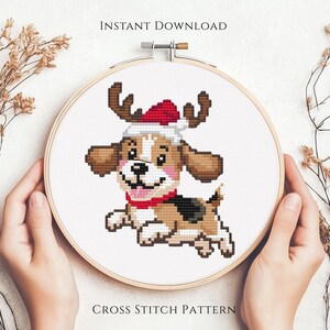 May include: A circular wooden embroidery hoop containing a cross stitch pattern of a brown and white puppy wearing a red and white Santa hat with reindeer antlers. The text INSTANT DOWNLOAD and CROSS STITCH PATTERN is printed on the image.
