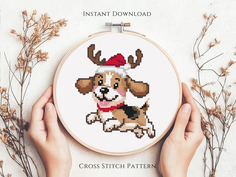 May include: A circular wooden embroidery hoop containing a cross stitch pattern of a brown and white puppy wearing a red and white Santa hat with reindeer antlers. The text INSTANT DOWNLOAD and CROSS STITCH PATTERN is printed on the image.