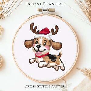May include: A cross stitch pattern in a wooden embroidery hoop featuring a cartoon puppy with brown and white fur, wearing a red and white Santa hat with brown reindeer antlers and a red collar, set against a plain white fabric background.
