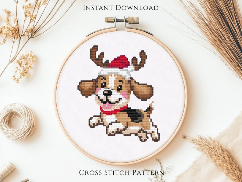 May include: A cross stitch pattern in a wooden embroidery hoop featuring a cartoon puppy with brown and white fur, wearing a red and white Santa hat with brown reindeer antlers and a red collar, set against a plain white fabric background.