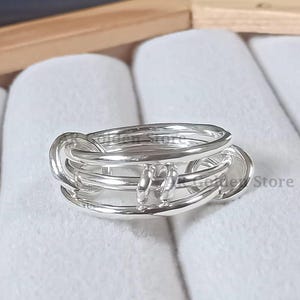 以下が含まれることがあります： ユニークなデザインのシルバーリング。リングは複数の絡み合ったバンドが特徴で、レイヤー効果を生み出しています。金属は研磨された反射面を持っています。リングは木箱の中の白い表面に展示されています。