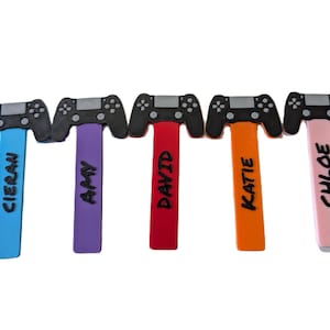 May include: Five colourful game controller-shaped bookmarks. Each bookmark has a black controller design at the top and a different coloured stem with a name printed on it: Cieran, Amy, David, Katie, and Chloe. The background is white.