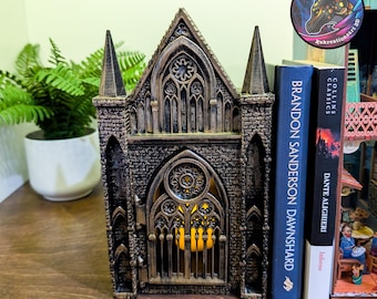 May include: A gothic-style decorative lantern with a warm, glowing interior. The lantern is dark gray with bronze accents, featuring intricate architectural details. Two books, "Brandon Sanderson Dawnshard" and "Inferno" are propped up against the lantern.