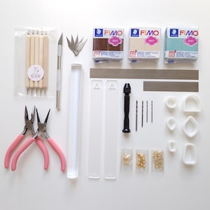 May include: A collection of craft supplies for polymer clay projects, including pink-handled pliers, clay tools, FIMO clay in brown, beige, and teal, a roller, cutters, a drill, and jewelry findings. The items are arranged on a white surface.