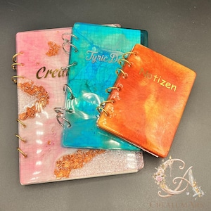 Puede incluir: Tres cuadernos de resina coloridos con encuadernación en espiral dorada. Los cuadernos son rosa, turquesa y naranja, con letras doradas que dicen "Create", "Tyric D" y "Notizen". El cuaderno rosa tiene detalles de purpurina dorada.